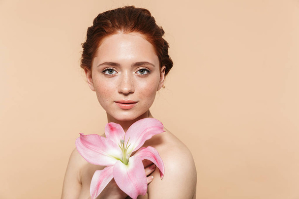 Pretty young redhead woman posing isolated over beige wall background pink flower. - Foto, Bild