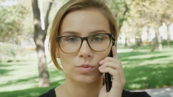Счастливая молодая белая женщина в очках разговаривает по черному телефону, сидя на скамейке в городском парке в летний день. Крупный план портретов
.  - Кадры, видео