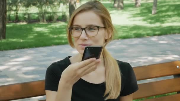 glücklich Mädchen in Brille mit einem Smartphone Spracherkennung Audio ai Nachricht Sprachfunktion online zu Fuß auf einer Straße mit Stadthintergrund, Sprachnachricht Helfer Studentin im Park auf Bank  - Filmmaterial, Video
