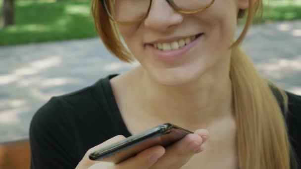 Chica feliz en gafas usando un teléfono inteligente reconocimiento de voz audio ai función de voz mensaje en línea caminando en una calle con fondo de la ciudad, ayudante de mensajes de voz mujer estudiante en el parque en el banco
  - Metraje, vídeo