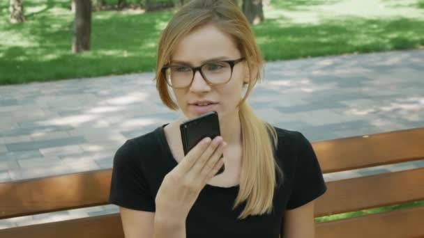 glücklich Mädchen in Brille mit einem Smartphone Spracherkennung Audio ai Nachricht Sprachfunktion online zu Fuß auf einer Straße mit Stadthintergrund, Sprachnachricht Helfer Studentin im Park auf Bank  - Filmmaterial, Video