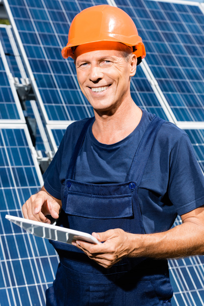 handsome engineer in t-shirt and orange hardhat smiling and holding digital tablet  - Фото, изображение