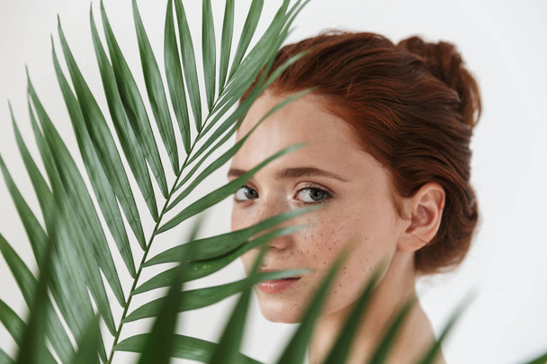 Young redhead woman posing isolated over white wall background with leaf green flowers. - Photo, image