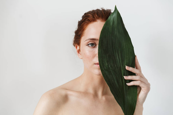 Young redhead woman posing isolated over white wall background with leaf green flowers. - Foto, Imagem