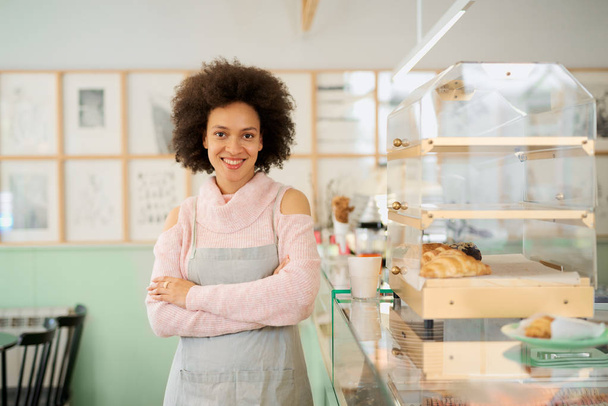Hermosa sonriente mestiza propietaria femenina de pastelería vestida de delantal de pie con los brazos cruzados en la tienda
. - Foto, Imagen