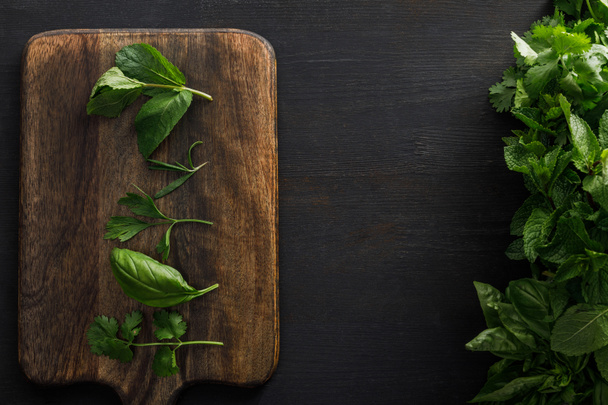 Top view of brown wooden cutting board with basil, parsley, cilantro and peppermint leaves near bundles of greenery on dark surface - Foto, immagini