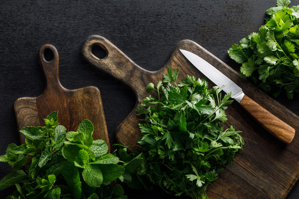Top view of brown wooden cutting boards with parsley, peppermint and cilantro  bundles with knife on dark surface - Photo, Image