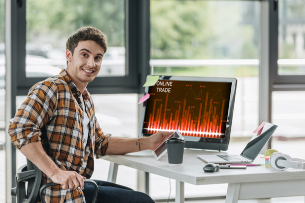 smiling programmer looking at camera while sitting near computer monitor with online trade on screen - Photo, Image