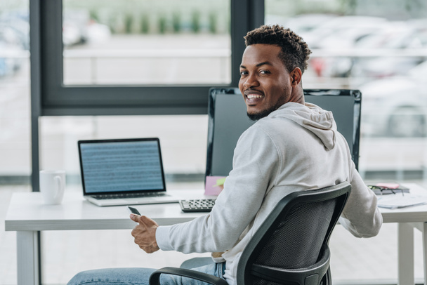 feliz afroamericano programador sonriendo a la cámara mientras está sentado en el lugar de trabajo en la oficina
 - Foto, imagen
