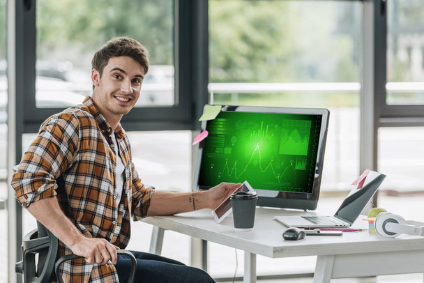 画面上のグラフやチャートでコンピュータモニターの近くに座っている間、陽気なプログラマーは、カメラで微笑んでいます - 写真・画像