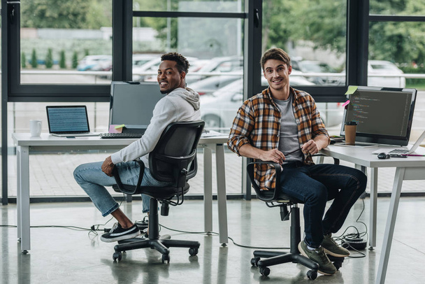 χαρούμενοι πολυπολιτισμικοί προγραμματιστές που χαμογελούν στην κάμερα ενώ κάθονται σε χώρους εργασίας - Φωτογραφία, εικόνα