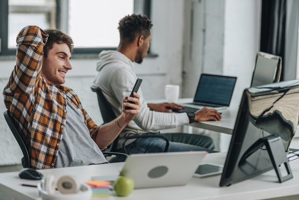 programador alegre usando el teléfono inteligente mientras está sentado en el lugar de trabajo cerca de un colega afroamericano
 - Foto, imagen
