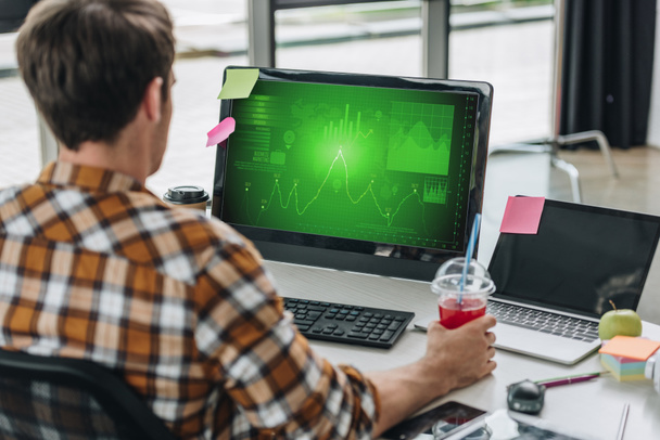 Widok z tyłu młody programista trzymając szklankę soku siedząc w miejscu pracy w pobliżu monitora komputerowego z wykresów i wykresów na ekranie - Zdjęcie, obraz