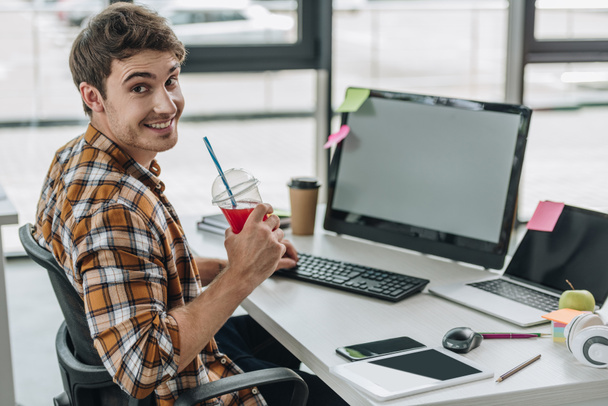 młody programista uśmiechając się do aparatu i trzymając szklankę soku siedząc w miejscu pracy  - Zdjęcie, obraz
