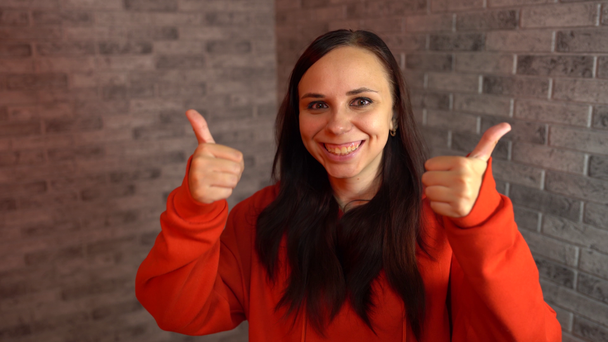 Een mooie emotionele vrouw in een rode hoodie lacht vrolijk en laat duimen zien. Een vrouw toont een gebaar van goedkeuring op een bakstenen achtergrond. Het concept van al dan niet leuk vinden. - Video