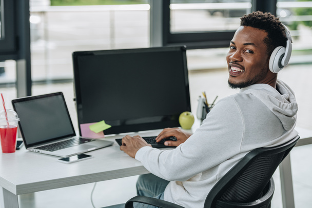 alegre afroamericano programador mirando cámara mientras sentado en el lugar de trabajo
 - Foto, imagen