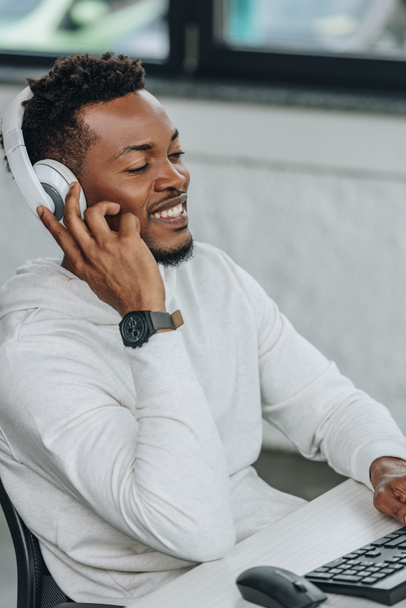 vrolijke Afro-Amerikaanse programmeur luisteren muziek in koptelefoon zittend op werkplek in kantoor - Foto, afbeelding