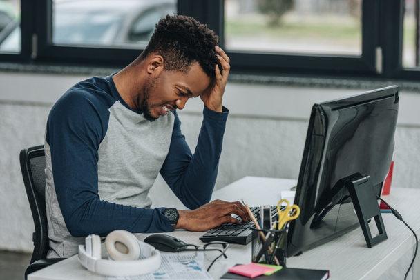upset african american programmer holding hand near head while working on computer in office - Photo, Image