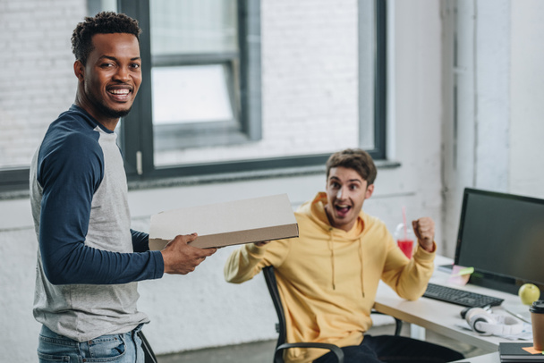 cheerful african american programmer holding pizza box while colleague showing yes gesture - Photo, Image