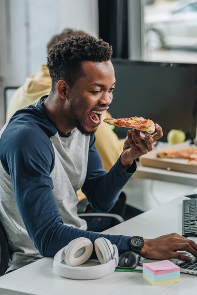 πεινασμένος αφρικανικός προγραμματιστής τρώγοντας πίτσα ενώ κάθεται στο χώρο εργασίας κοντά σε συνάδελφο - Φωτογραφία, εικόνα