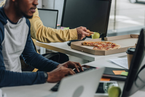 bijgesneden beeld van programmeur Holding stukje pizza in de buurt van African American collega - Foto, afbeelding