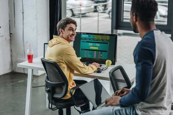 KYIV, UKRAINE - 29 de julho de 2019: programador sorridente usando o computador com o site Sportsbet na tela enquanto seu colega afro-americano sentado na mesa com laptop
 - Foto, Imagem