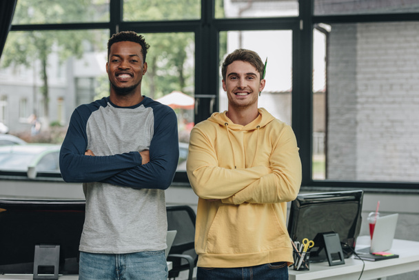 два веселых мультикультурных программиста стоят со скрещенными руками и улыбаются в офисе перед камерой
 - Фото, изображение