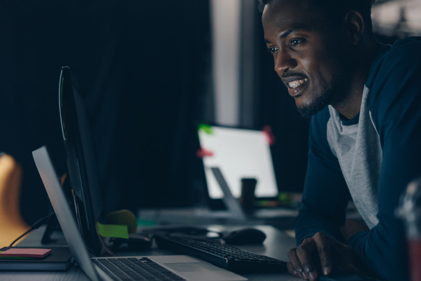 young african american programmer smiling while looking at computer monitor at night in office - Photo, Image