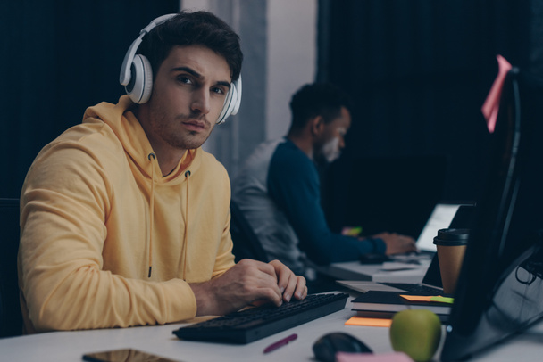 selective focus of young programmer in headphones looking at camera while working near african american colleague - Photo, Image
