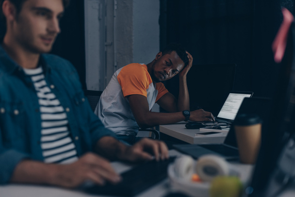 selektiver Fokus eines afrikanisch-amerikanischen Programmierers auf einen multikulturellen Kollegen, während er nachts im Büro zusammenarbeitet - Foto, Bild
