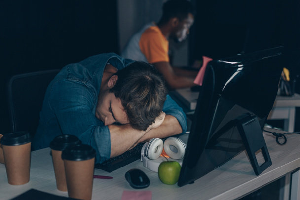 selective focus of tired programmer sleeping at workplace near african american colleague - Foto, imagen