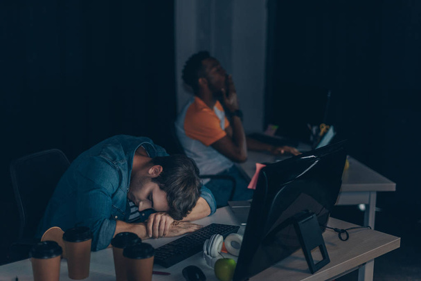 wyczerpany programista śpi w miejscu pracy, podczas gdy afrykański kolega pracujący w nocy w biurze - Zdjęcie, obraz