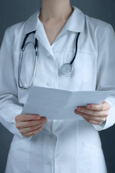 Caucasian girl in medical white clothes with a stethoscope closeup. Holding a sheet of paper in his hands. - Foto, afbeelding