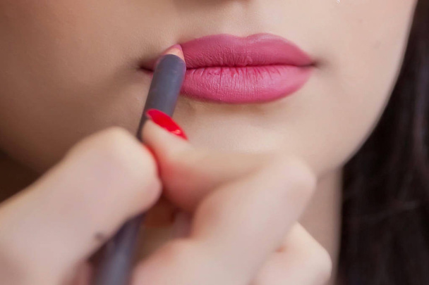 The female hand of visagiste is holding a pencil and drawing lips . Professional Makeup artist applies pencil lipstick bridal . Close up of unrecognizable pretty girl getting her lips painted. - Foto, afbeelding
