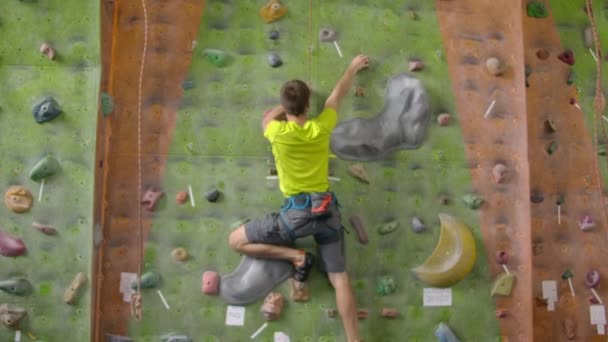 Male climber climbs the cliff wall in the room reaching and gripping hold. A professional concentrated climber moves up the wall, the camera rises and lays down his movements. Boom follow camera - Footage, Video