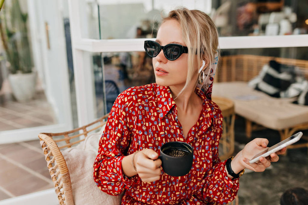 attraktive, stylische Kaukasierin mit schwarzer Sonnenbrille, die Kaffee trinkt und mit dem Smartphone spricht  - Foto, Bild