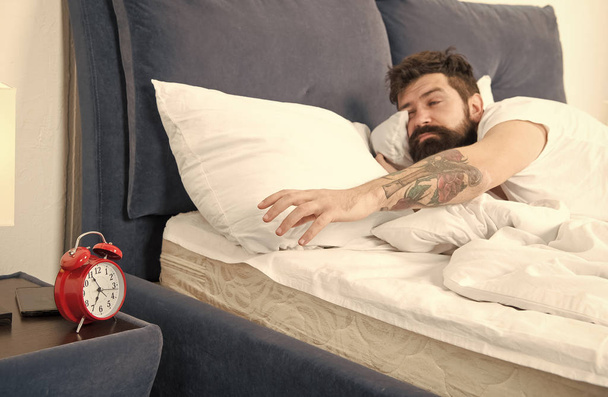 Overslept again. Tips for waking up early. Man bearded hipster sleepy face bed with alarm clock. Turn off that ringing. What terrible noise. Problem early morning awakening. Get up with alarm clock - Fotó, kép