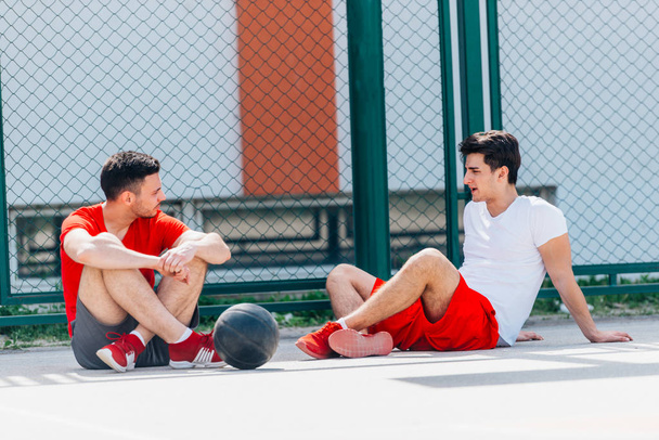 Two strong caucasian athletes resting on the ground at the basketball court while having a conversation. - Photo, Image