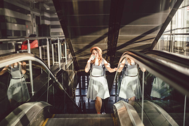 Retrato sonriente mujer con sombrero en el aeropuerto en escaleras mecánicas. personas que viajan con equipaje de mano. Turismo temático y transporte. Chica caucásica con café en la terminal del aeropuerto en una escalera mecánica
 - Foto, imagen