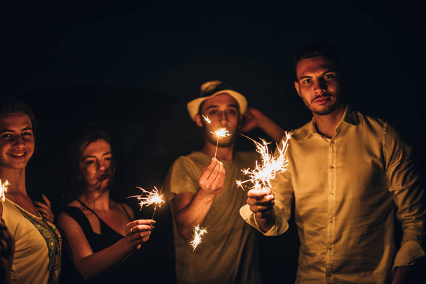 Group of friends lighting fire sparkle sticks in the dark and en. - Foto, Imagen