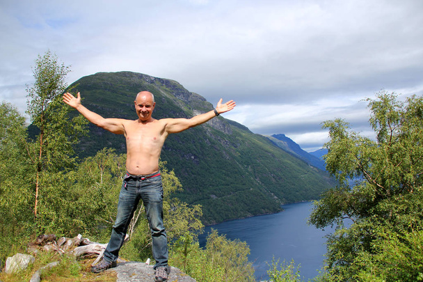 a man stands high on a mountain top above the fjord holding his hands up admiring the magnificent landscape - Foto, afbeelding