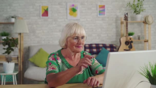 Thoughtful older woman finds a solution and uses a laptop sitting at a table in the living room - Záběry, video