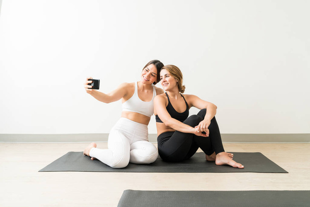 Smiling young woman taking selfie with friend sitting on yoga mat against wall - Photo, Image