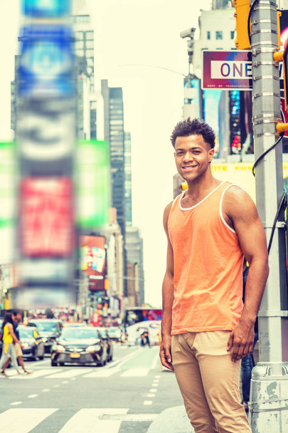 Молодой счастливый смешанный гоночный американец путешествует по Нью-Йорку жарким летом в оранжевой майке, бежевых штанах, стоит на пересечении улиц на Таймс-сквер Манхэттена, смотрит, улыбаясь
 - Фото, изображение