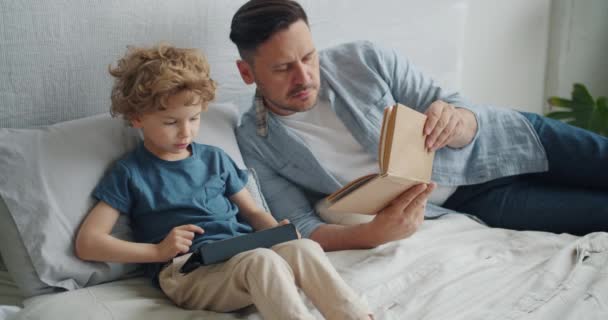 Father son spending day together reading book watching cartoons on laptop in bed - Imágenes, Vídeo