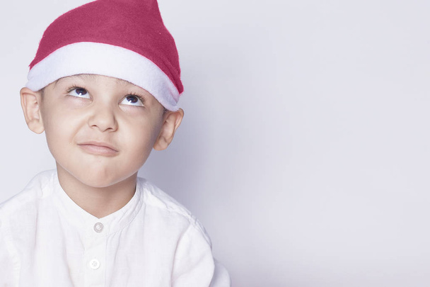 Όμορφο παιδί με το καπέλο του Αϊ Βασίλη που εύχεται ή ονειρεύεται κάτι. Το παιδί ονειρεύεται το χριστουγεννιάτικο δώρο. Το παιδί ψάχνει και σκέφτεται - Φωτογραφία, εικόνα