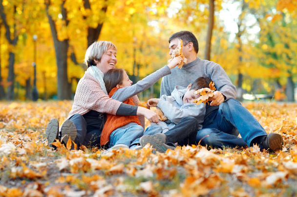 落ち葉の上に座って、遊んで、秋の都市公園で楽しんで幸せな家族。子供と親が一緒に素敵な一日を過ごします。木々の明るい日差しと黄色い葉、秋の季節. - 写真・画像