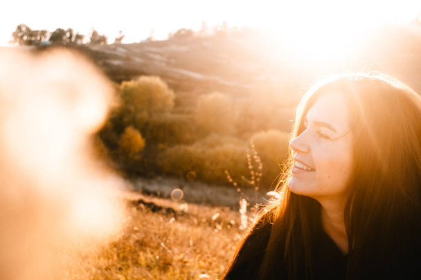 Portrét nádherné brunetky mladé ženy v černé kožené bundě a košili při západu slunce na pozadí kopců. Efekt odlesku objektivu. Kopírovat místo - Fotografie, Obrázek