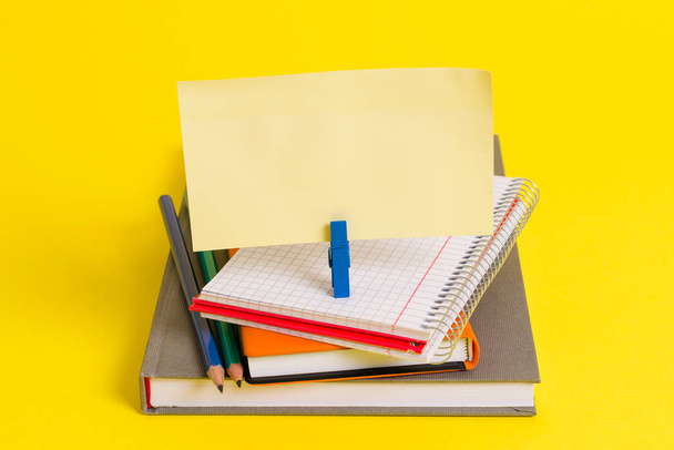 Books, pencils and an empty rectangle square shaped reminder above a blank opened notebook held by a blue clothespin. Office supplies in a yellow background. - Photo, Image