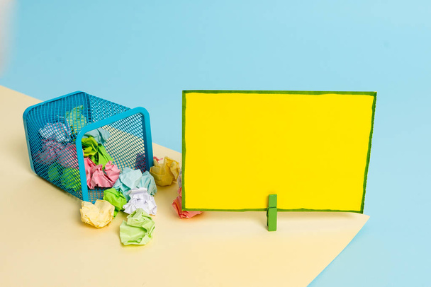 Μικρό κάδο σκουπιδιών γεμάτο από τσαλακωμένο χαρτί και ένα μανταματάκι που κρέμονται μια πολύχρωμη χάρτινη νότα σε κίτρινο και μπλε φόντο. Αναλώσιμα γραφείου και κενή υπενθύμιση. - Φωτογραφία, εικόνα